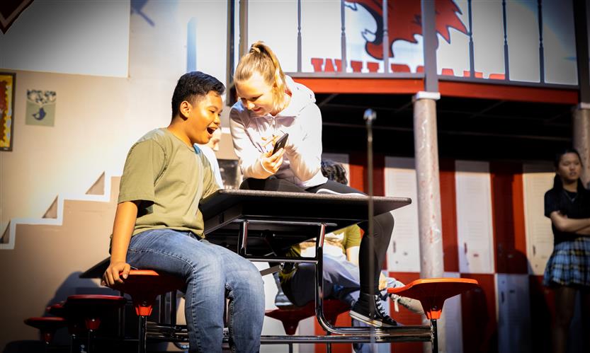 A student playing a character sits on a lunch table onstage